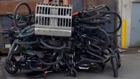 Multiple confiscated e-bikes being crushed. A pile of bikes can be seen with a digger bucket on top of them. 