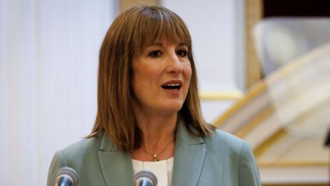 Chancellor Rachel Reeves wearing a sage green jacket and white collarless blouse stands behind a podium with two microphones giving a speech at the annual Mansion House dinner in the City of London, UK.