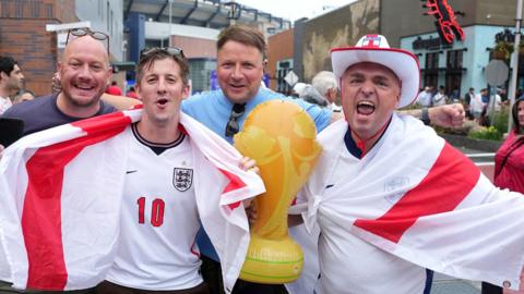Four male England football fans draped in England flags outside Boston Football stadium. They have an inflatable  world cup trophy with them.