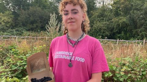 Alex Vardill wearing a pink T-shirt, smiling straight at the camera and holding a box of blackberries.