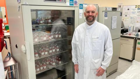 David Usherwood standing in the laboratory. He is in front of a temperature controlled cupboard, in which corneas are kept. You cannot see the corners in the image, but each is contained inside a small labelled bottle filled with reddish orange fluid.