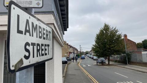 A view of Lambert Street. A sign bearing the road's name is on the left and, in the background, there is a row of houses and cars.