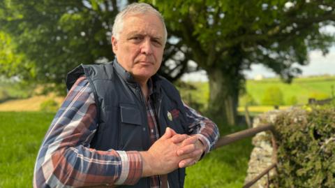William is standing in a grassy field and is leaning over a metal gate. He is wearing a colourful plaid shirt and navy gilet. He has grey hair.