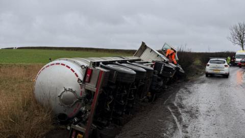 A HGV tanker overturned on a country road. The vehicle has fallen on its side into a ditch and has flattened vegetation. Other vehicles can be seen further along the road. A person in an orange hi-vis top and bottoms is perching on a front wheel looking into the cab. 