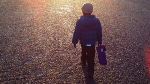 A boy in a blue jacket and school uniform, holding a blue book bag and wearing a hat, walks across tarmac