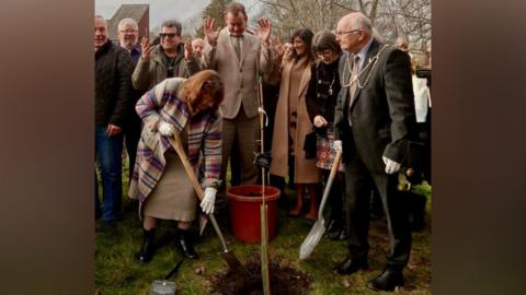 A group of people plant a tree including, on the right, a man in a suit and mayoral chains