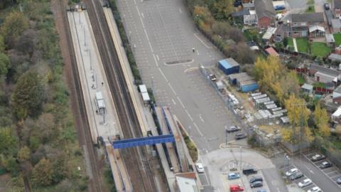 Ariel view of Swinton railway station in Rotherham. A blue footbridge is crossing the tracks between platforms. The station car park is also in view, with a majority of the parking spaces empty. Nearby houses can also be seen. On the other side of the railway line are trees