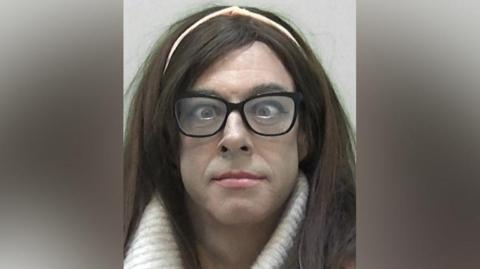 Mugshot of Natalie Wolf, formerly known as Ryan Haley, who has thick long brown hair with a pink hairband in, with slight make up and pink lipstick and black-framed glasses over a pair of cross-eyed eyes.