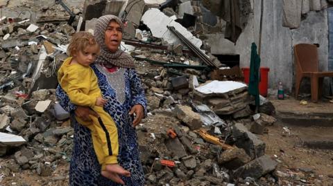 A Palestinian woman holds a child dressed in a yellow track suit, with a pile of rubble behind them.