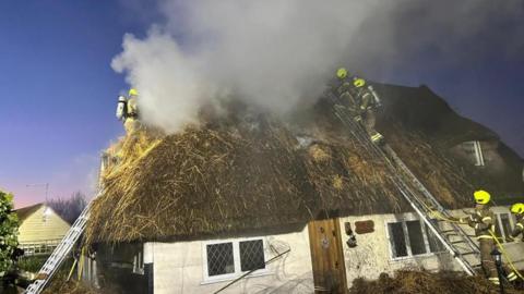 A white cottage with a thatched roof which is covered in smoke. Three firefighters are on the roof and two further ones are on the ground. 