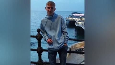 A young man wearing a blue zip up hoody is stood with an elbow leaning on a railing with the sea and a pier behind him.
