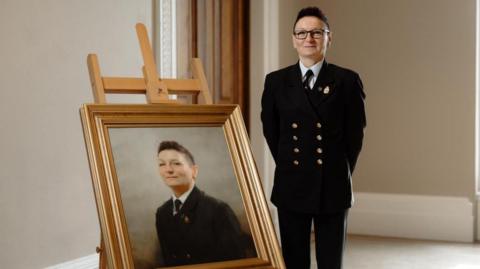 Karolina Jabryzk, wearing a formal dark uniform with brass buttons, stands beside an easel displaying a framed portrait of her, set against a neutral background. The setting is a room with light-coloured walls.