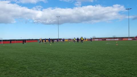 A football pitch with a number of players doing training drills. There are small cones on the grass and some of the people are wearing yellow training bibs. The scene shows one half of the pitch from halfway line to goal. There is a low red wall surrounding the playing area with a number of advertising boards attached. Two floodlights tower above the far touchline. The sky is blue with a long white cloud.