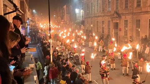 A torchlit procession of people all wearing striped tops. There are crowds along the edge of the street and on balconies above the street.