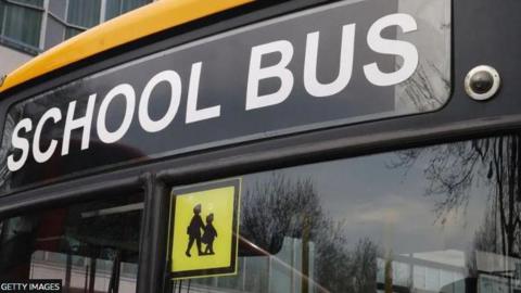 Image shows the front windscreen of a school bus with the words school bus written at the top of the windscreen and in the middle of the windscreen at the top is a shadow picture of two children with a yellow background. 