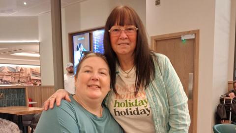 Rebecca is sat on the left of the photo and her mum Delia is stood to her right with her arm around her. Rebecca is wearing a blue t-shirt. Delia is wearing glasses and a stripy shirt over a white graphic t-shirt which says 'holiday' four times.