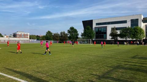 Two women's football teams, wearing red and pink kits, playing a match on an outdoor pitch. A black and white college building is in the background behind some trees.