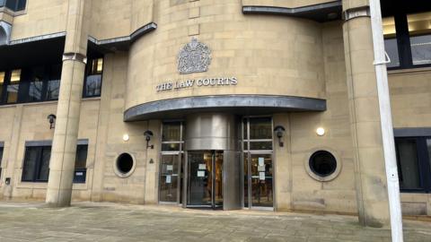 The front of Bradford Crown Court, a multi-storey building made of sand-coloured brick. The words "The Law Courts" as well as a crest can be seen above the main entrance.