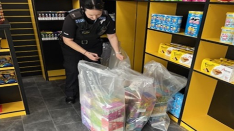 A female police officer is holding several plastic bags inside one of the shops which are filled with vapes and tobacco. The bags are on a grey floor next to yellow shelving displaying goods