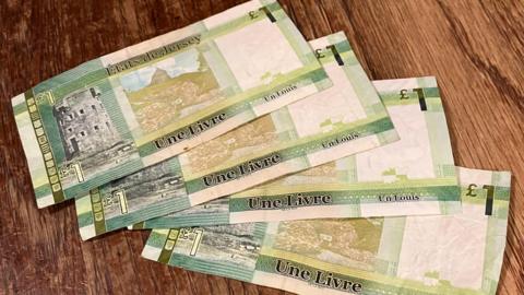 Four Jersey £1 notes have been placed on a wooden table. The notes are green and white.
