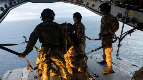 Military personnel at the edge of a plane, wearing combat fatigues - they are over a body of water and preparing items to be airdropped