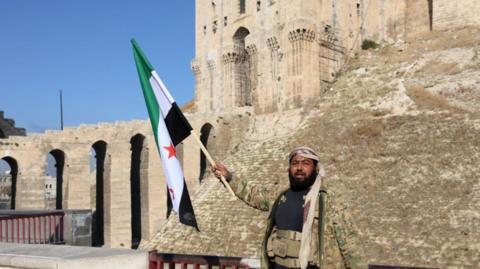 A man in a camouflage jacket stands in front of Aleppo's citadel. He is waving an opposition flag with his right arm.