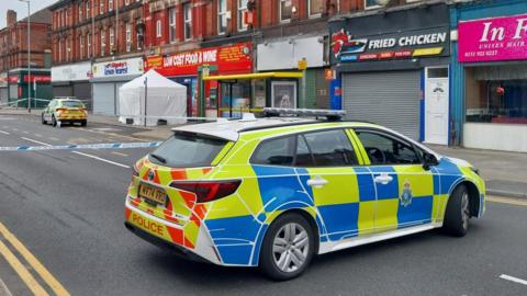 A yellow and blue liveried police car outside a police cordon tape stretched across a street. Shops line the street at the back and beneath red brick properties. Another police car is in the background.