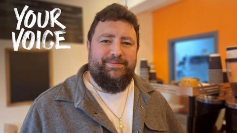 Daniel King is standing in a cafe with a coffee machine and counter behind him. He is smiling slightly into camera. He has short brown hair and a beard. He wears a white t shirt and grey shirt with a silver cross on a chain around his neck.