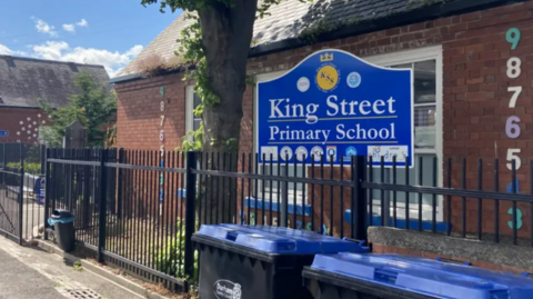 The exterior of King Street Primary School, which sits behind a fence and has a blue sign with the school's name prominently displayed and colourful numbers on the walls.