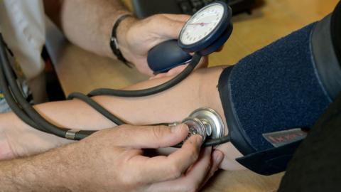 The arm of someone having their blood pressure checked at a desk. There is a blue Velcro band around the patient's upper arm and a doctor is using a stethoscope and monitor to check the blood pressure. 