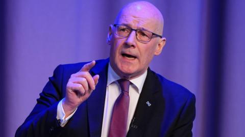 John Swinney, who is bald and is wearing glasses, is wearing a blue suit, white shirt and purple tie and is sitting in front of a purple background. He has a saltire tie-pin on his left lapel.