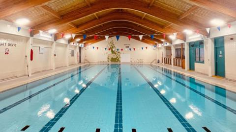A brightly lit swimming pool covered by a wooden roof. The pool is blue and surrounded by white tiles. Flags are draped across the water at the top and bottom of the facility.