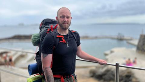 A bald, bearded man in his late 20s by some railings in front of some outdoor bathing pools. He has tattoos on his forearm, wears a black T-shirt and green shorts, and carries a large backpack.