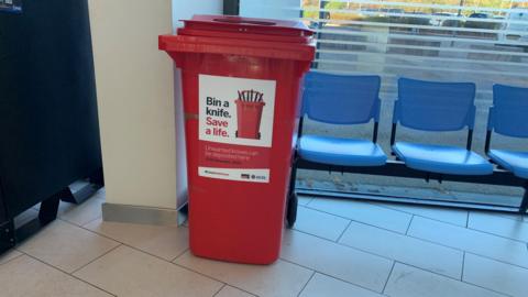 A red wheelie bin with a sign on it reading Bin a knife, Save a Life.