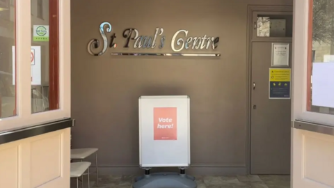 A photo of the foyer of St Paul's Centre. There is a sign with the venue name and an advertising board with a 'Vote Here!' poster attached to it. 