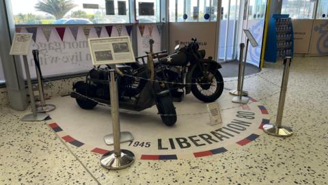 A display with a barbed wire barrier. There a two motobikes - a Harley-Davidson WLA (on the right) and a Cushman Airborne Scooter. There are Signs with information on. On the floor it says "1945 LIBERATION 80 2025". There is red, blue and white bunting behind. 