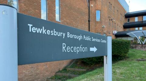 Large three story brick building with a sign in front reading Tewkesbury Borough Public Services Centre. Underneath is the word Reception and an arrow pointing you to the door.