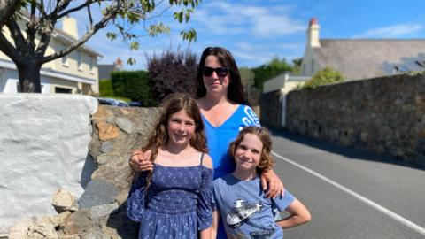 Claire with her two children stood on a pavement. A road is on the right. The woman has sunglasses on and dark hair. They are all smiling towards the camera.