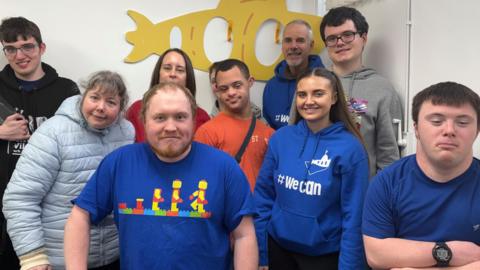 Ten people stand in the corner of a white room with a cut out of a yellow submarine behind them. From left to right, stands a young man who is a member of the group wearing a black hoodie, holding a black tablet which helps him communicate. He has dark brown hair and wears glasses. Next is a woman with grey hair who wears a pale blue puffer coat. The lady next to her has dark brown hair with a red t-shirt on, she works for the charity. There is then two young men, one wearing a grey hoodie and the other an orange t-shirt. There is then another member of the charity. He stands in the back corner and has grey hair. He wears a staff hoodie with the words 'we can' on. There is then a tall young man wearing a grey hoodie and black framed glasses. At the front of the picture is a young woman and two young men- all wearing navy blue and smiling at the camera.