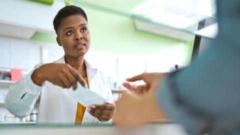Stock photo shows a pharmacist holding a box of medication as she hands it over to a patient who is out of view, in a pharmacy setting.