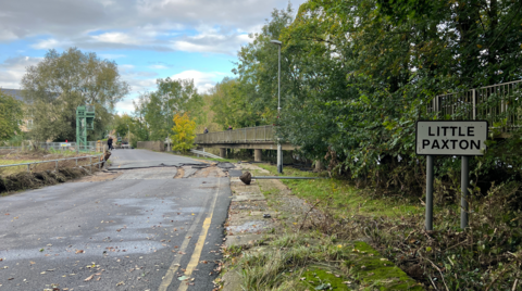 The left side of the photo shows Mill Lane stretching into the distance. There are chunks of tarmac missing in the centre of the road. The right side features a "Little Paxton" sign and a metal post that has been pulled from the ground during the floods. The sky is blue with some clouds and there is greenery and trees on both the right and left sides of the road.  