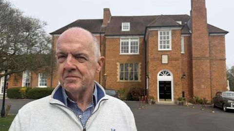 A man wearing a blue checked shirt with a pale grey jumper over the top stands at the forefront of the picture, looking off to the left of the camera. Behind him is a red brick building, with tall windows and a black door.