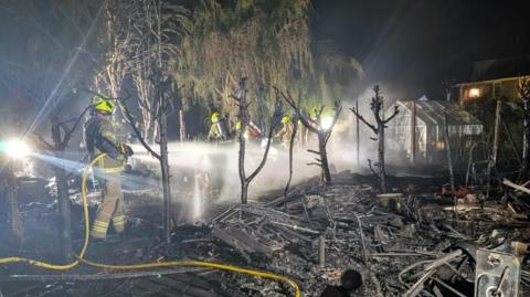 Firefighters hose down a burnt and destroyed garden. Trees can be seen burnt with rubble strewn across the garden. 