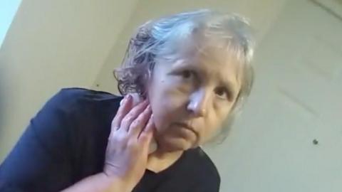 A screengrab of police body worn video showing Stefania Glowka stood in a corridor holding a bandage or tissue to a wound on her neck. She is a woman in her 60s with grey hair and is wearing a dark top.