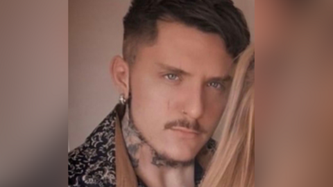 A young man with styled dark hair, cropped at the sides, is wearing a brown and black patterned top with a turned down collar. He has a trimmed moustache and beard, a silver earring and tattoos on his neck.