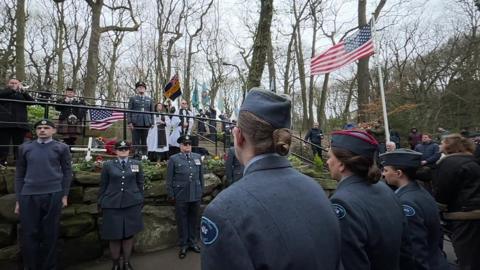 Members of the British and American military gather for a memorial service in a park - flags are flying including the stars and stripes