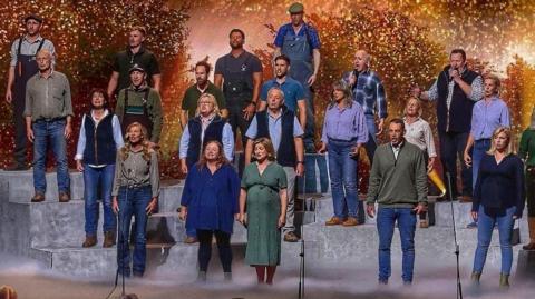 A large group of performers stands on tiered platforms on a brightly lit stage, appearing to sing together as a choir. A conductor stands at the front, guiding the performance. In front of the stage, a panel of judges sits at a table, watching. The background features warm, glowing lights and foliage-like scenery, while an audience in the foreground raises hands and watches the show.