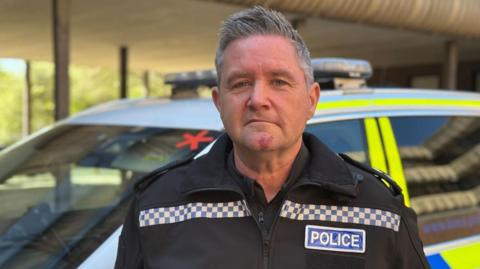 A man with short grey hair that is styled in an upward fashion is wearing a black jacket and shirt with 'POLICE' in blue and white on the side. He is pulling a serious expression into the camera and behind him is a white, yellow and blue police car parked up that is blurred.