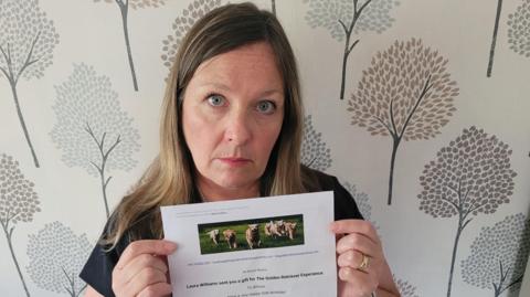 Women with long hair. She's looking sad to the camera. She's holding an A4 paper document with a voucher with dogs running around 