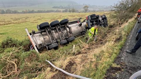 A tanker on its side in a field in a very rural location.  A pipe is being used to drain its contents to another lorry.